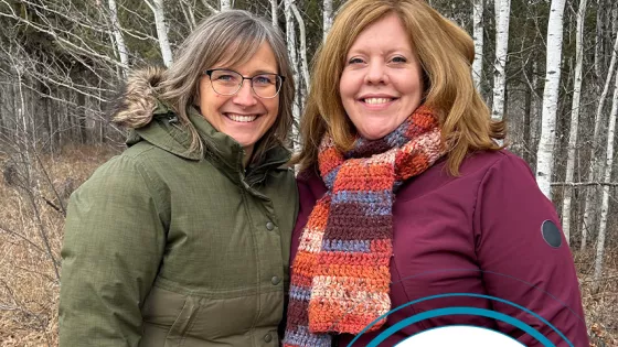 Two women standing in a forest and smiling at the camera.