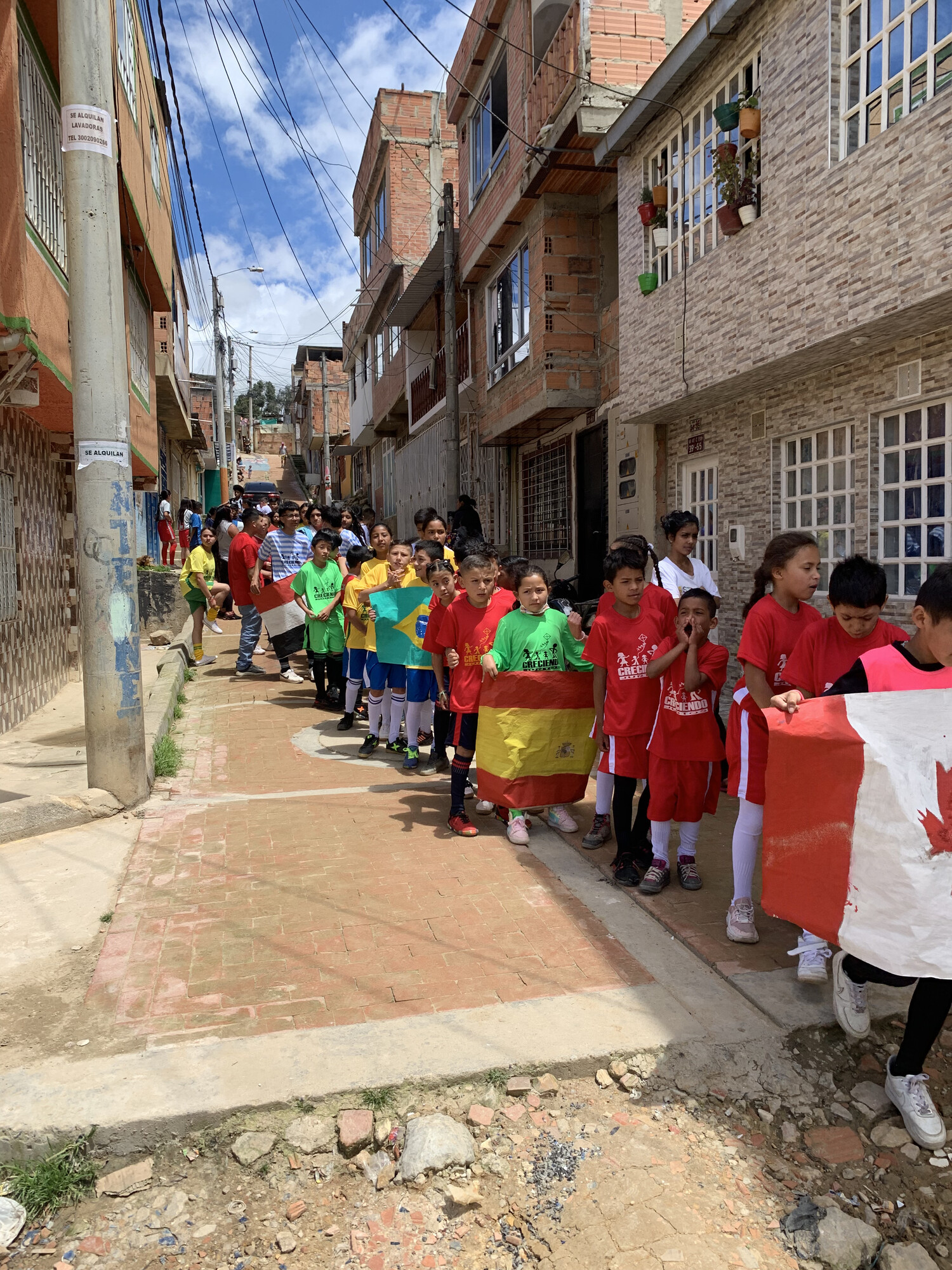 Children stand in a long line on a narrow street in Colombia. A few of them are holding different country flags.
