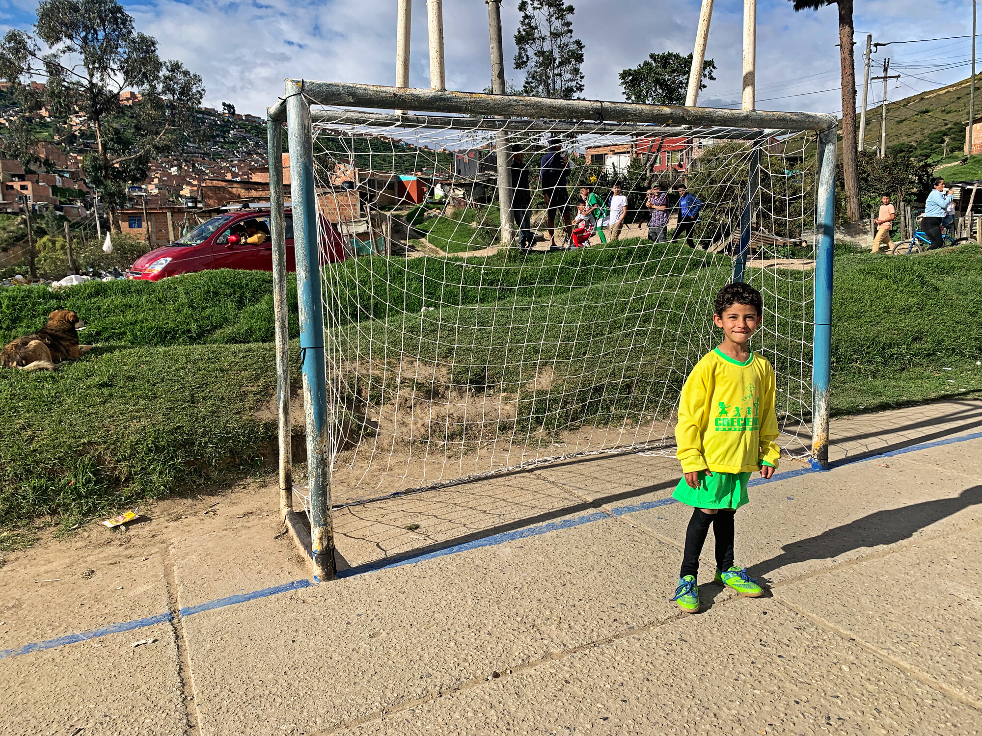 A child, wearing a yellow and green soccer uniform, stands in an empty goal.