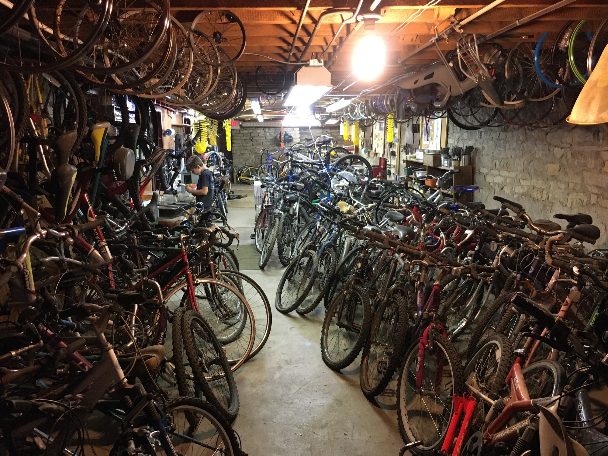 A storage room full of bikes. In the background, a kid works on a repair.
