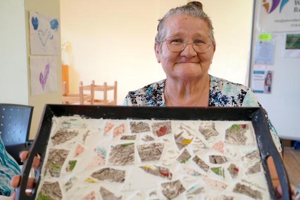 An elderly woman holds up a mosaic tray