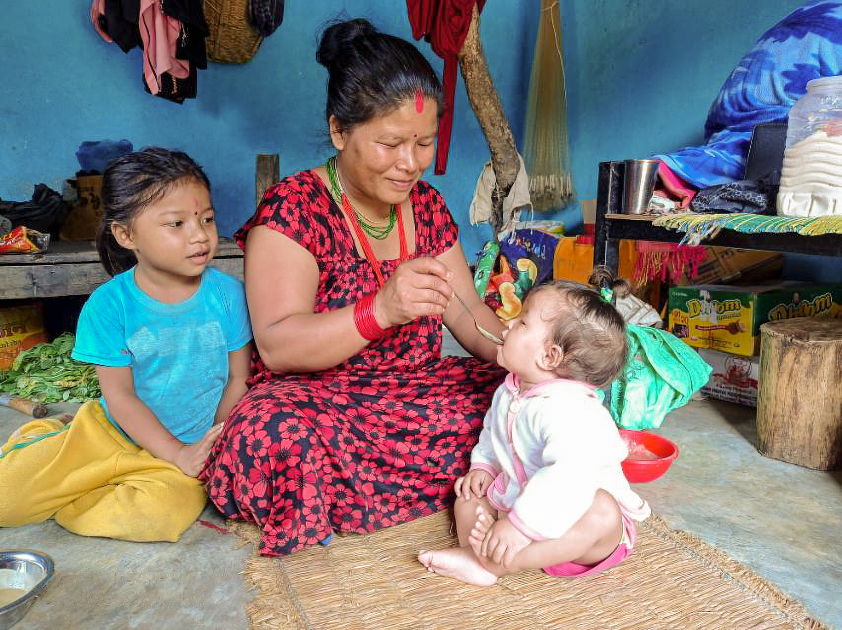 A Nepali woman sits on the ground and feeds a baby with a spoon. Another child is sitting to the left of her.