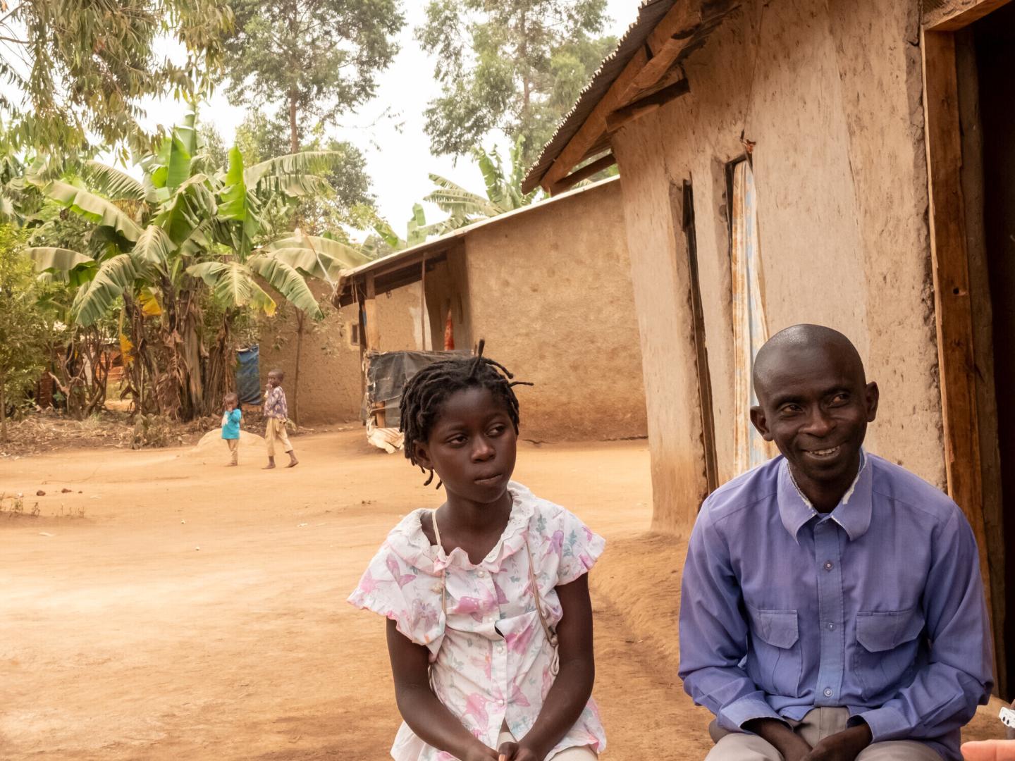 A Ugandan father and daughter sit next to one another outside their home