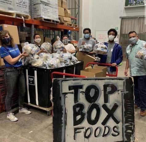 Six young adults stand in warehouse holding plastic bags of food. There is a sign in front of them that says, "Top box foods."