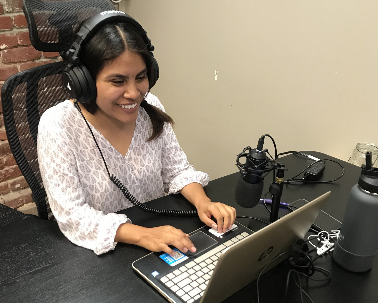 A young woman with podcasting gear sits at a table with a laptop.