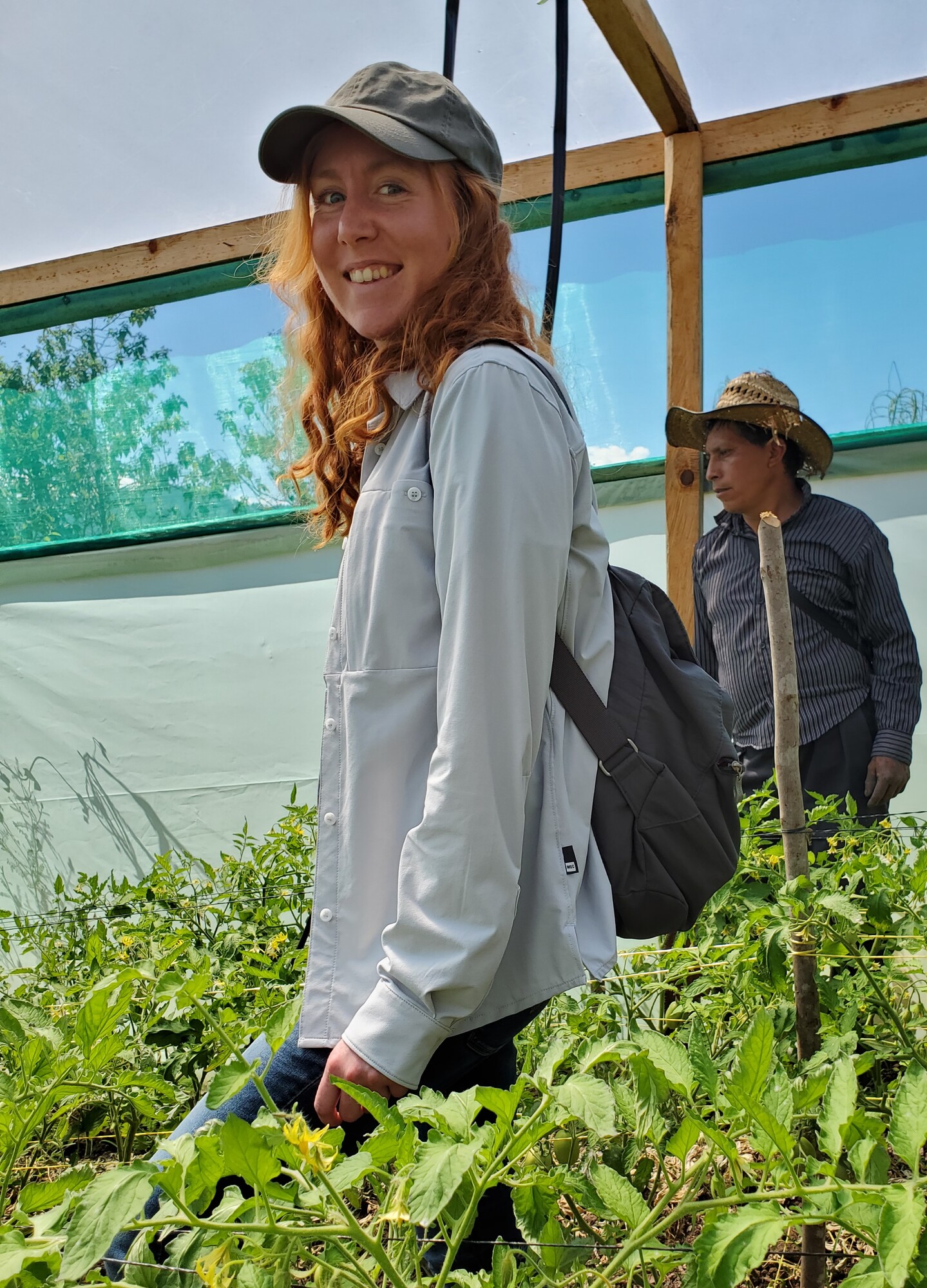 A young woman with red hair and a ball cap stands in a greenhouse surrounded by green leafy plants
