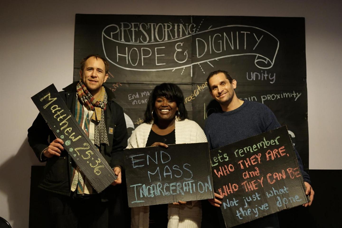 A group of three people pose in a makeshift photo booth with signs that say, "Matthew 25:26," "End mass incarceration" and "Let's remember who they are and who they can be not just what they've done."