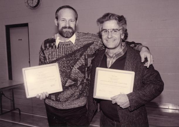 Two men stand together holding certificates. One has his arm around the other's shoulders.