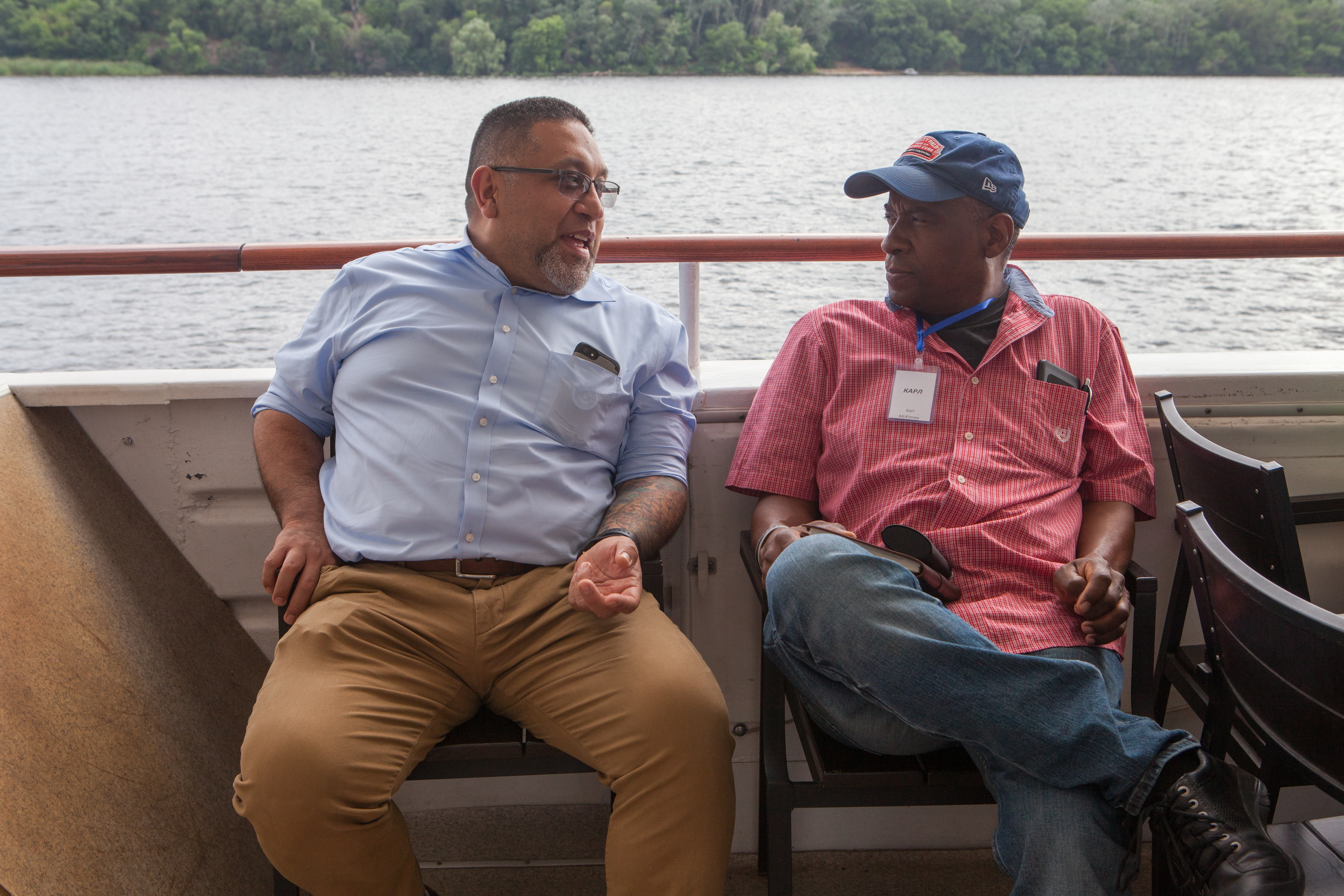 Two men sit near the railing of a boat. They are talking to each other.