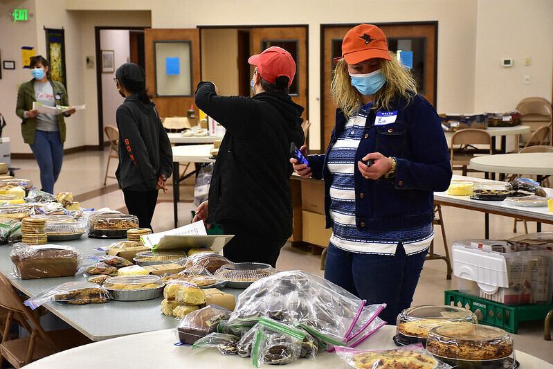 A woman in a ball cap and a face mask stands in front of a table full of baked goods