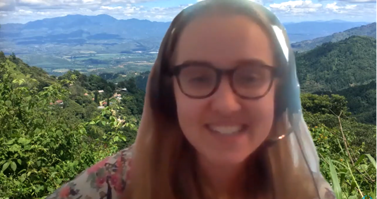 A young woman speaking during a webinar zoom call. There is a zoom background of mountains behind her.