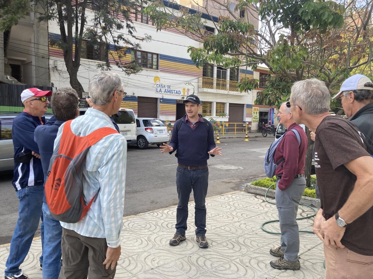 A man in a ball cap talking with a group of people on the side of a street