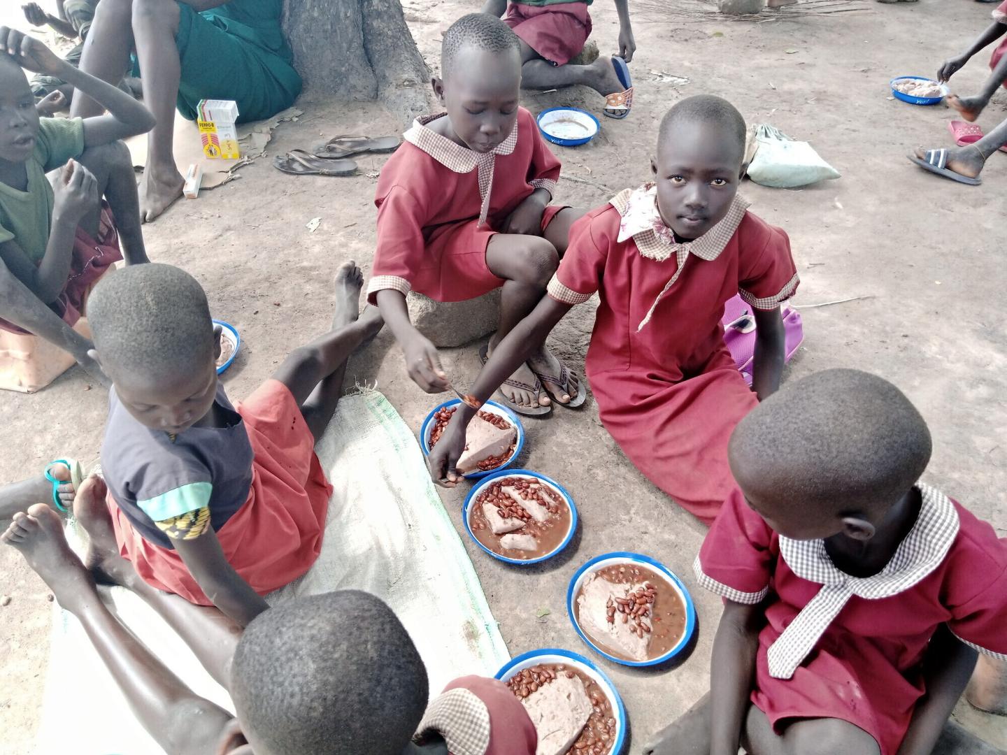 Five South Sudanese students in red uniforms sit on the ground and eat from plates