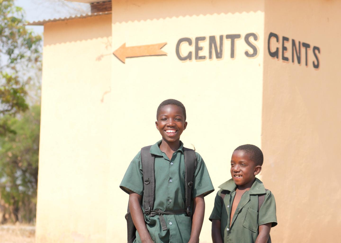 Two students in green stand in front of a latrine that says "Gents"