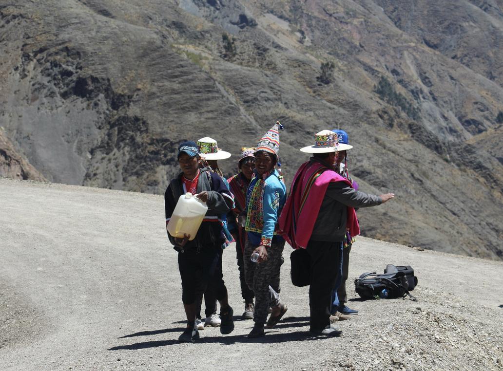 A group of Bolivians stand on the side of a road. One man is holding a large container of tea