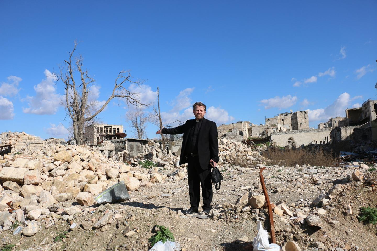 A Reverend in a clerical collar stands on a pile of rubble in Syria