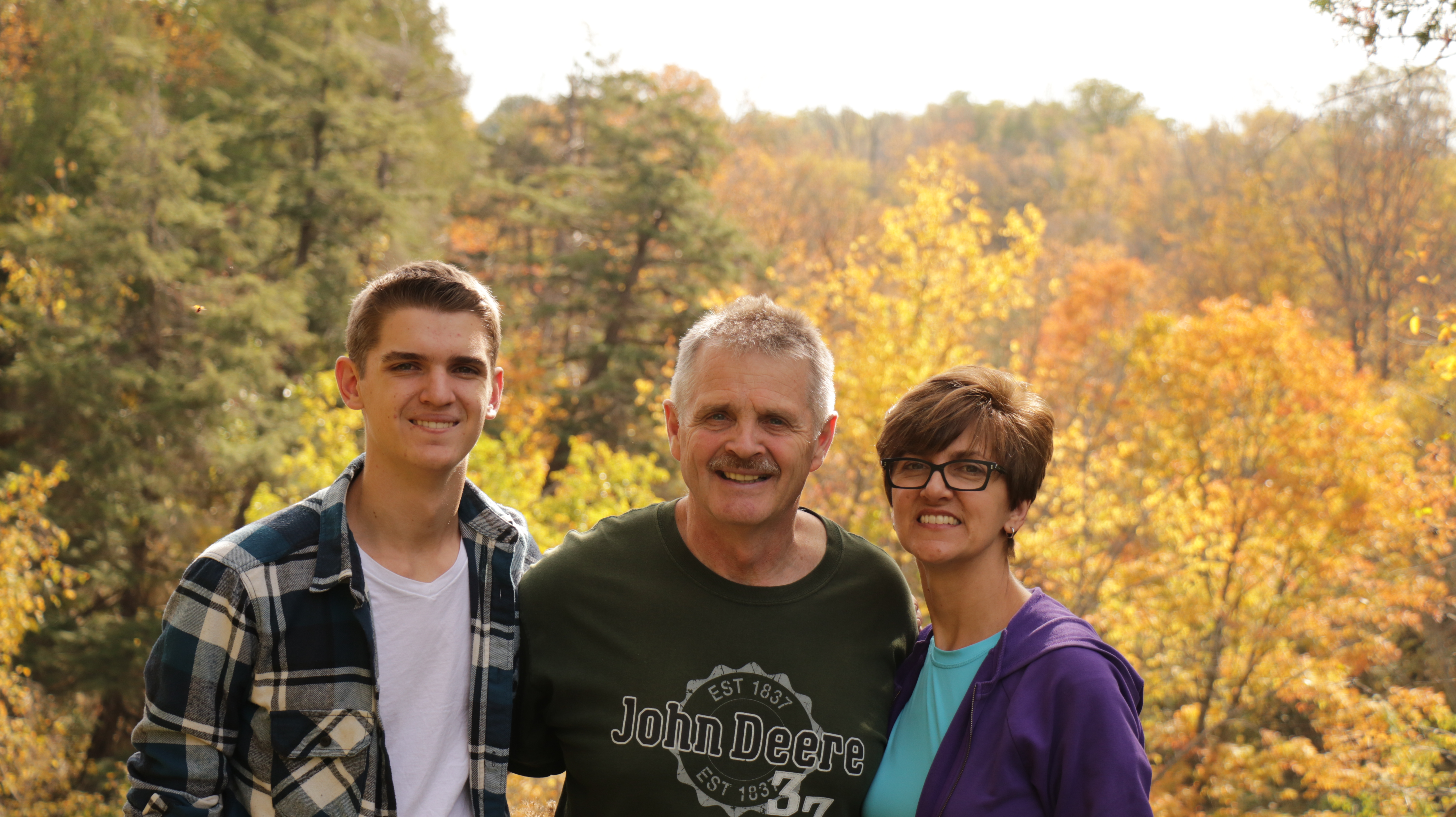 A young man, a middle-aged man and middle-aged woman smile at camera with a forest behind them.