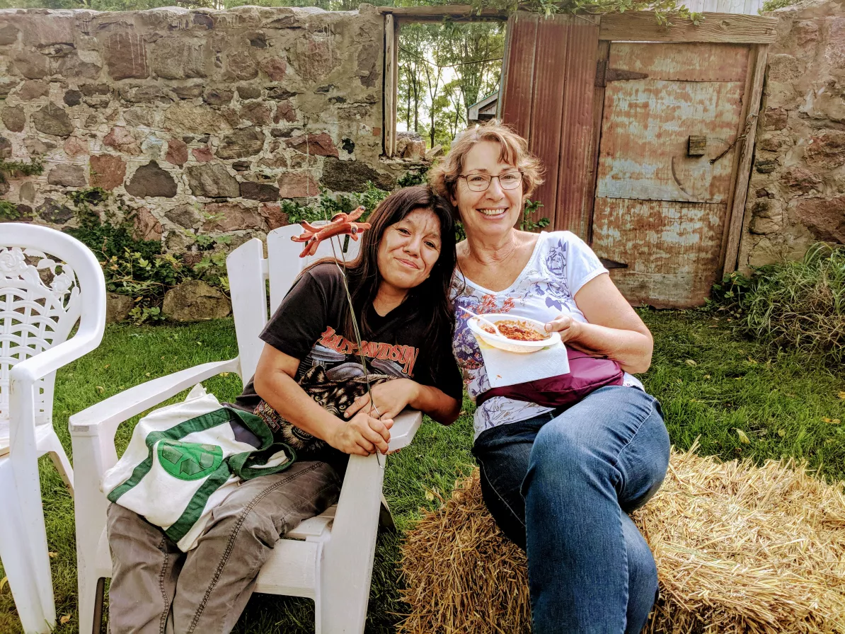 Two woman sit next to each other. The woman on the left is sitting in a white lawn chair, holding a hot dog on a stick and is leaning into the woman on the right. The woman on the right is sitting on a hay bale