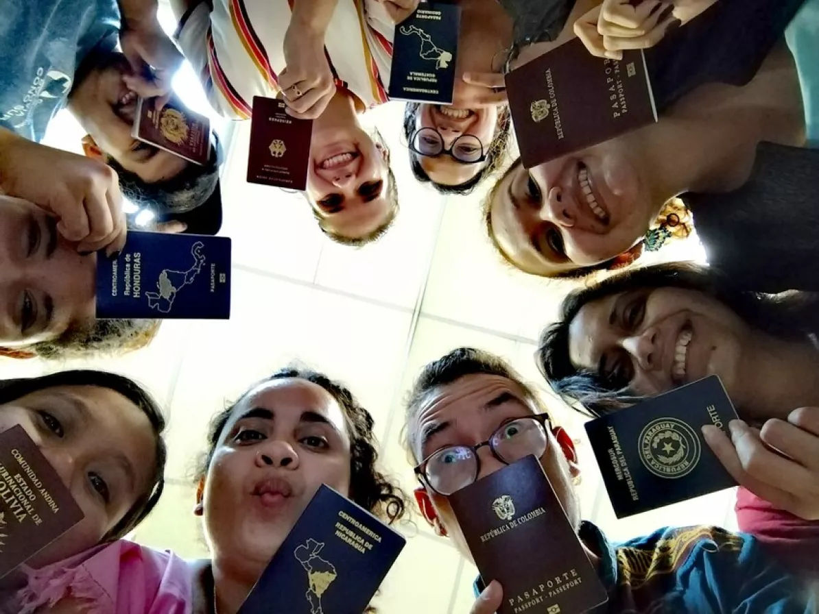 Nine young adults of different nationalities stand in a circle and look down at the camera. They are holding passports.