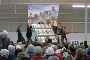 Three people display a quilt during an auction. Behind them is an auctioneer.