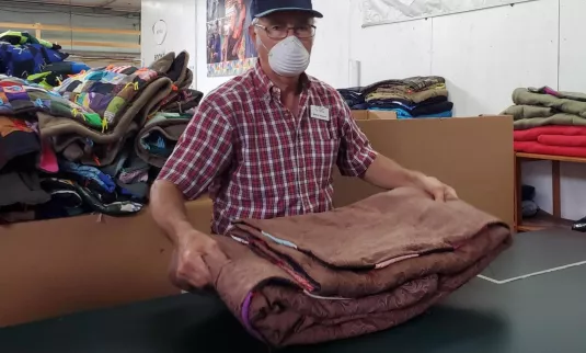 A man in a ball cap and a face mask holds a folded comforter in a warehouse.