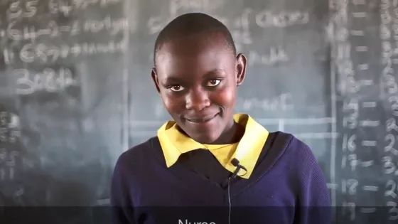 A young Kenyan student stands in front of a chalkboard. She is wearing a school uniform sweater with a yellow collar.