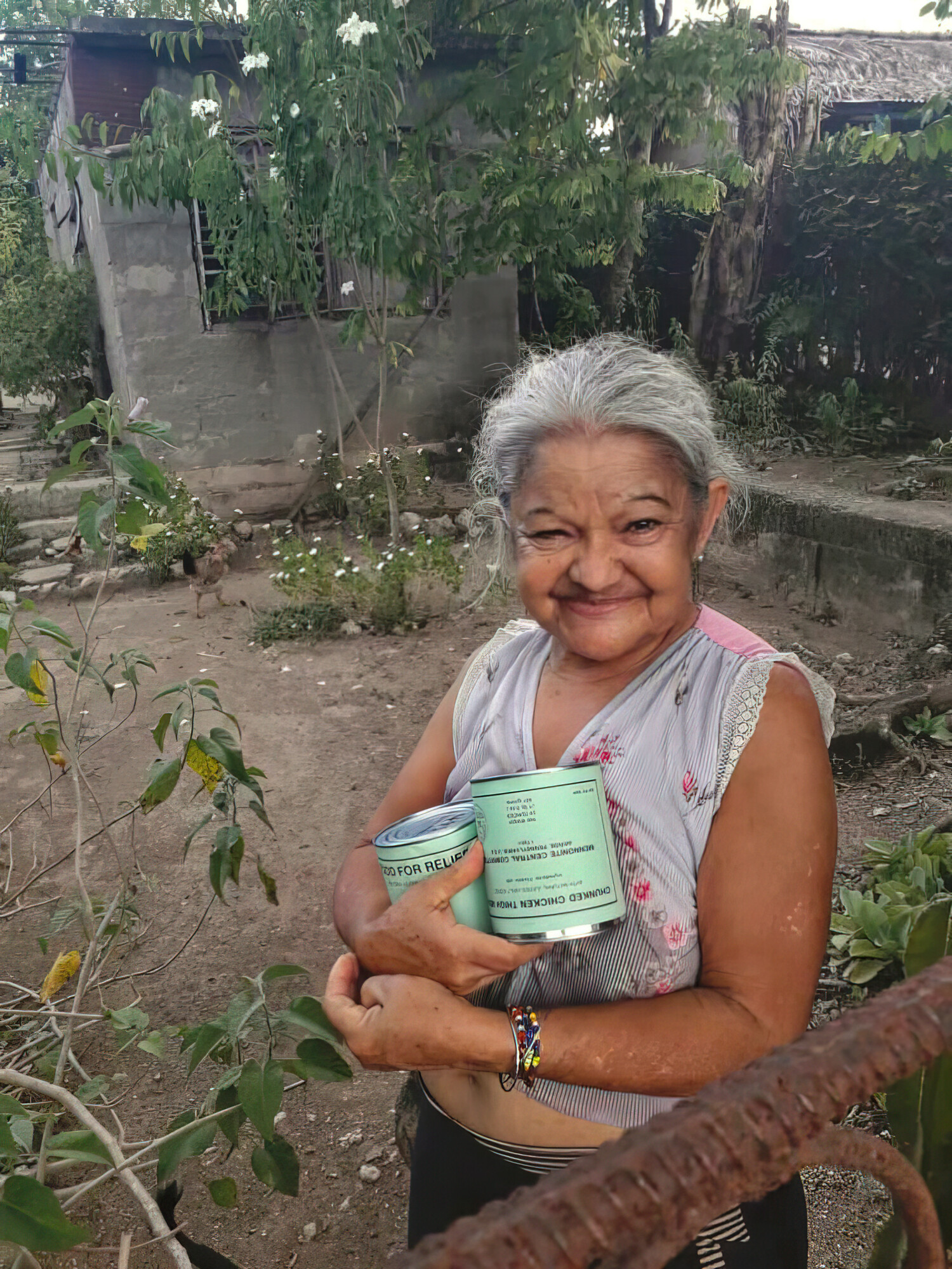 An older Cuban woman stands in a yard and holds two cans of meat with green labels.