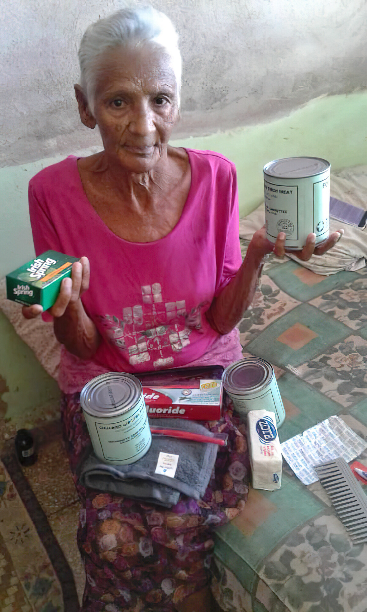 An older Cuban woman sits on a bed. She is holding relief supplies. There are also relief supplies resting on her lap.
