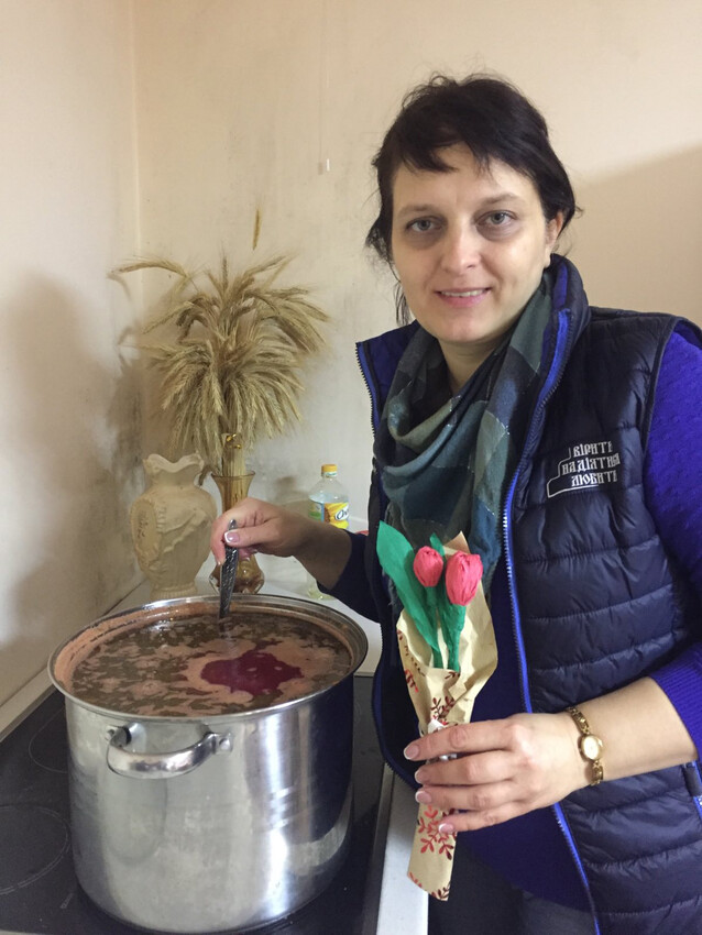 A Ukrainian woman stands over a large soup pot. She stirs the pot with a spoon in one hand. She is holding a small bouquet of roses in her other hand.