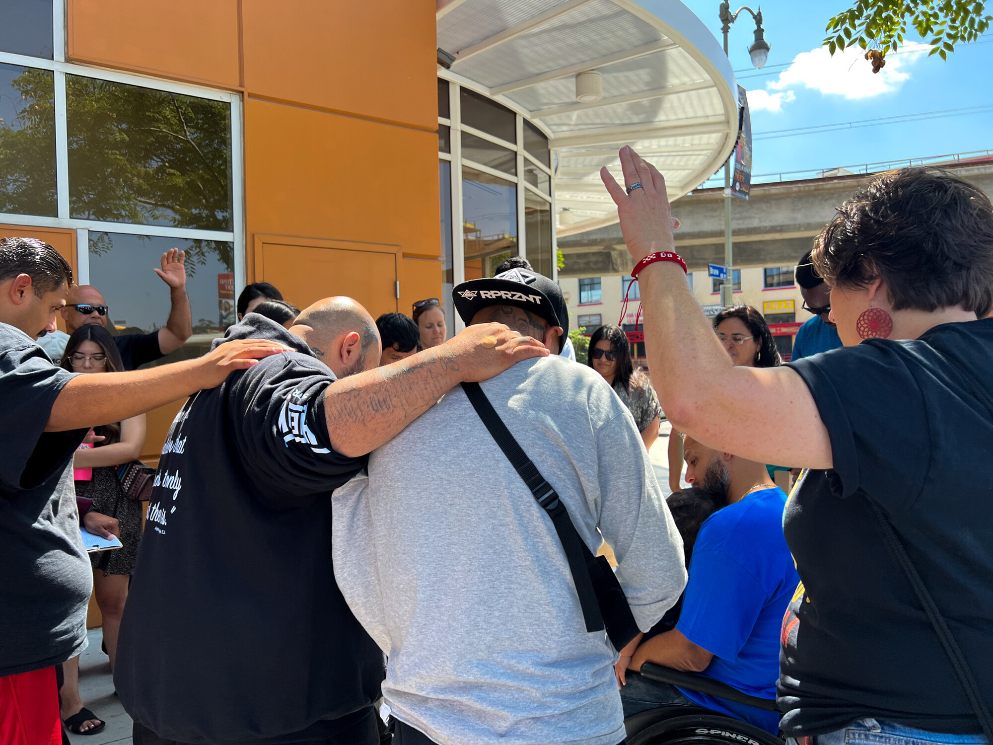 A group of young adults stand in a group with their arms around each other, praying