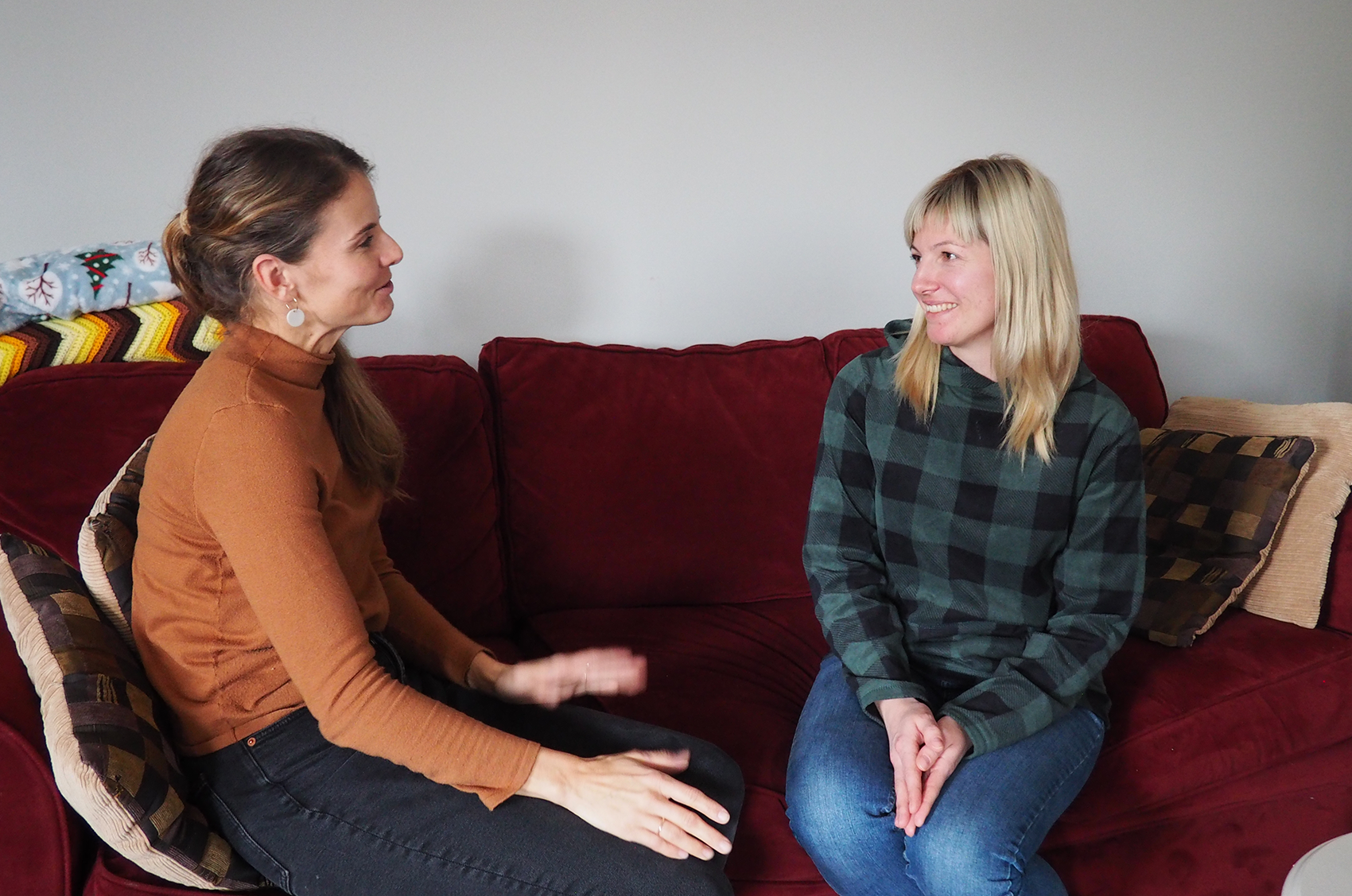 Two women sitting on a red couch having a discussion. The woman on the left is speaking, while the woman on the right smiles and listens.