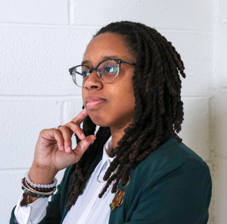 A woman with long dreads and glasses with her hand up to her chin in a thinking pose