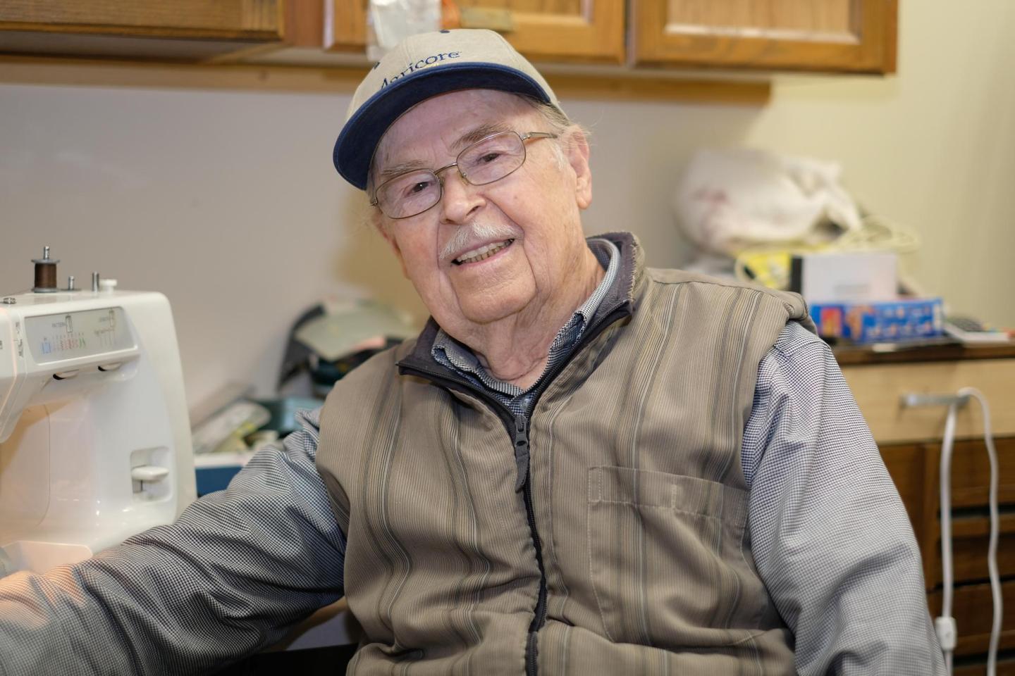 An older man in a hat and glasses sits by a sewing machine