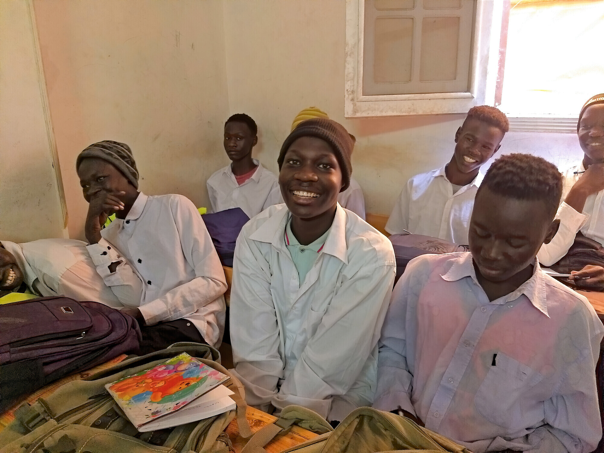A group of Sudanese students sit at their desks in a small classroom.