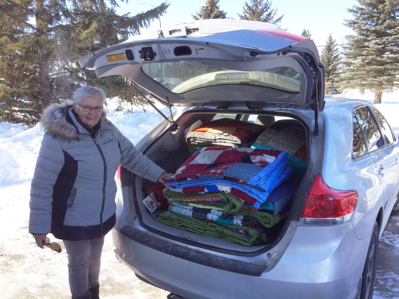 A woman in a winter coat stands by the open truck of a hatchback car. Inside the car are piles of colorful comforters.
