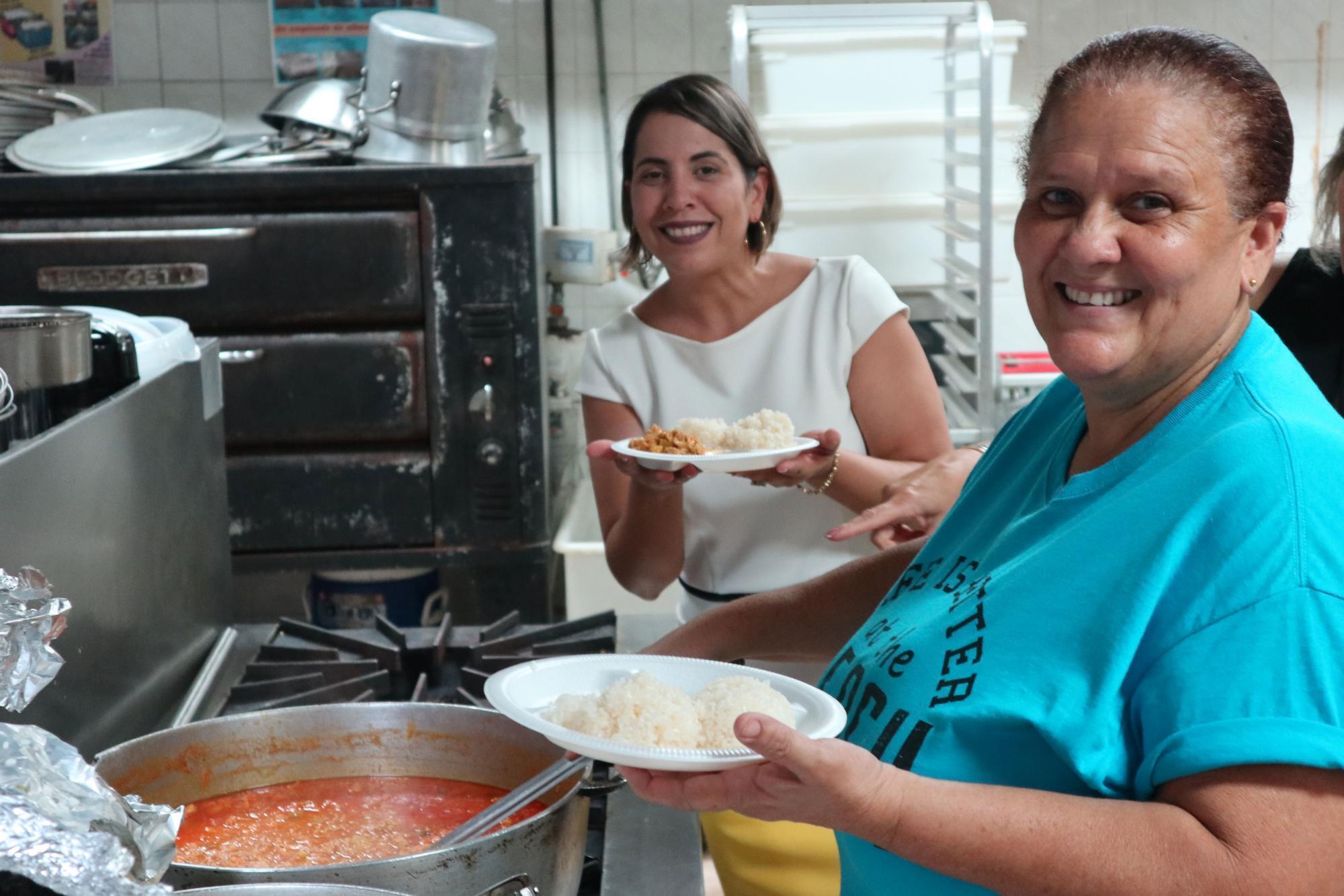 Two women hold plates of rice over a large pot of beans cooking on an industrial sized stove