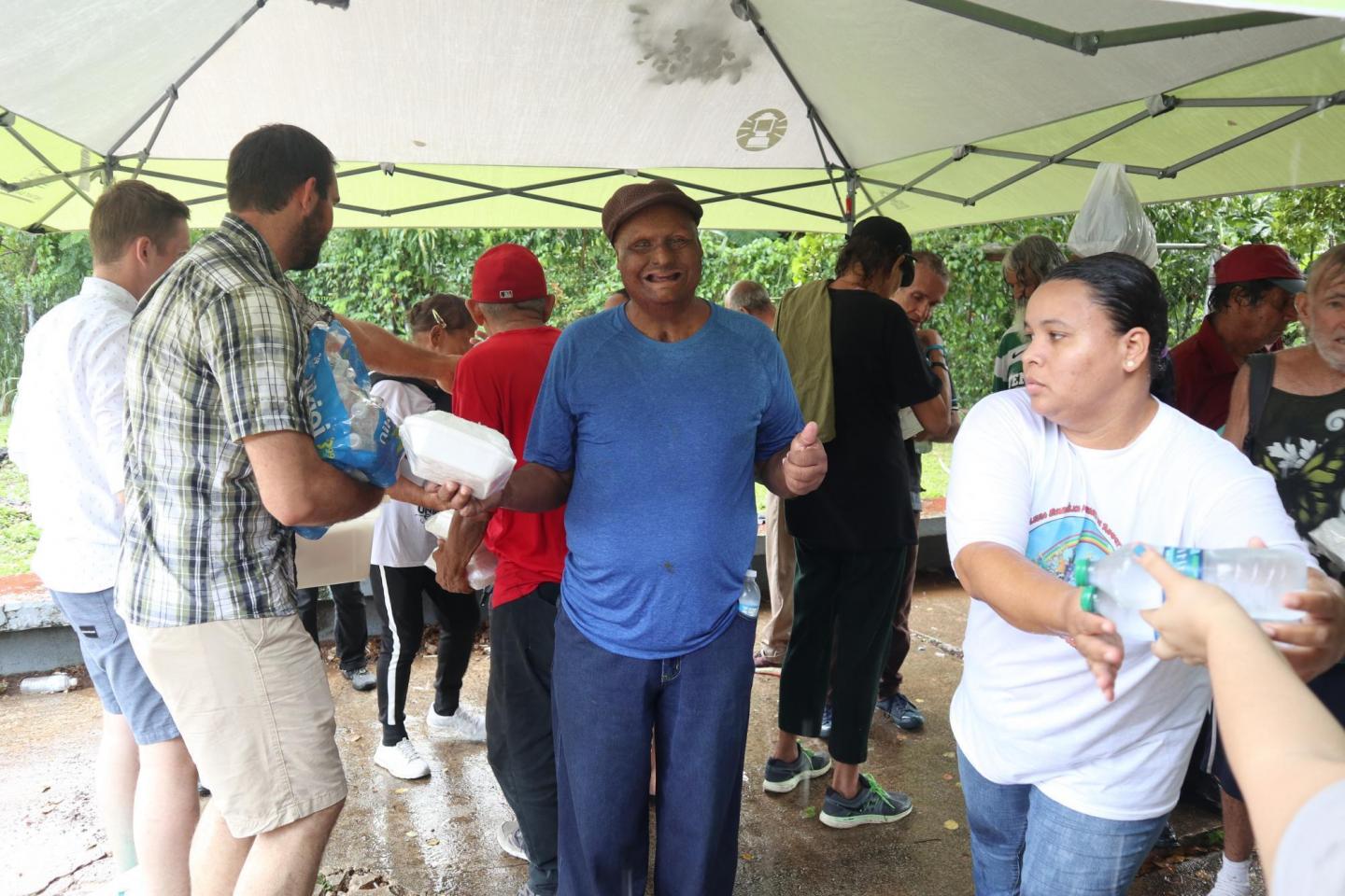 In the midst of a group of people, a man in a blue shirt smiles and gives a thumbs up. He is holding a to-do food container. 