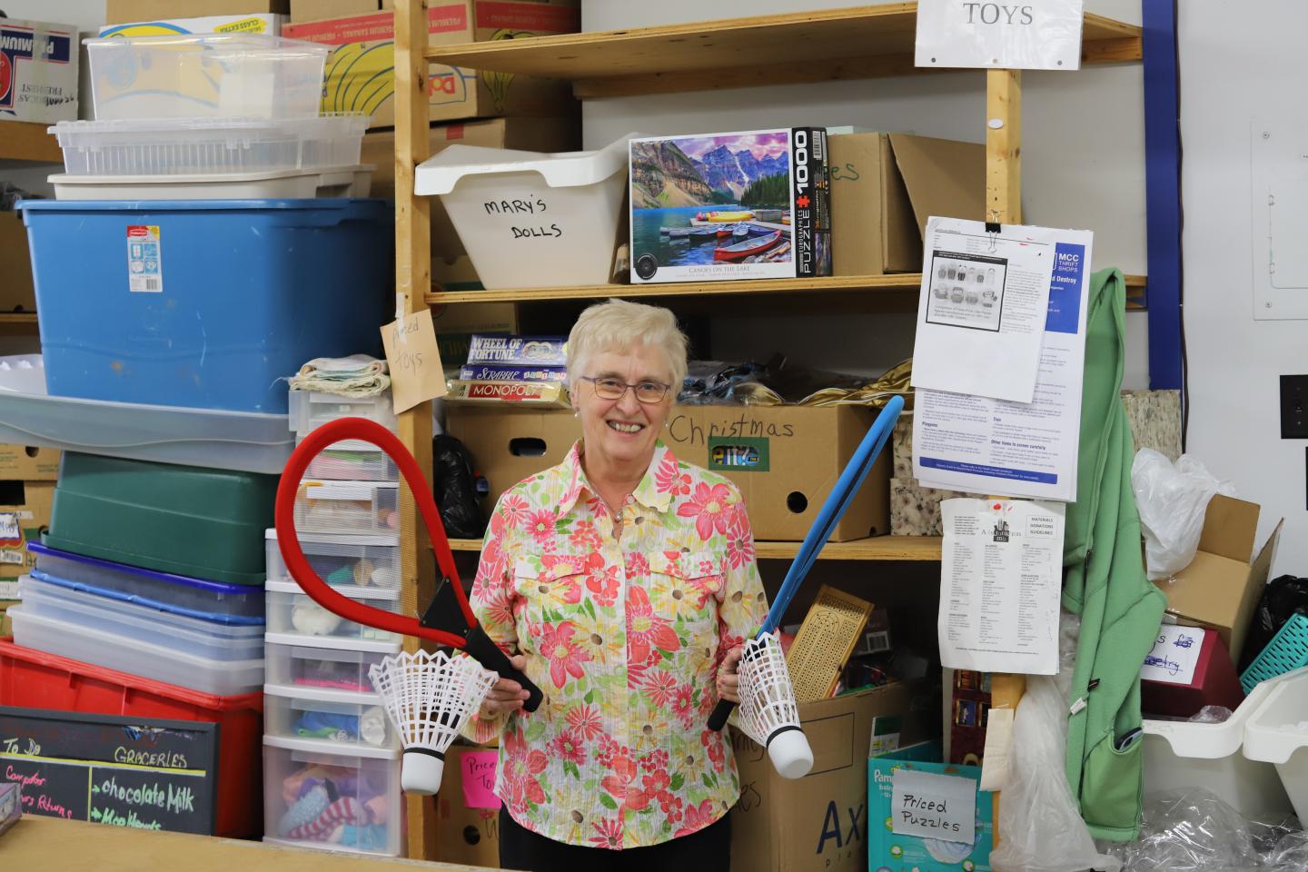 An older woman in a floral shirt stands in front of a wall of thrift shop donations. She is holding large badminton rackets and birdies 