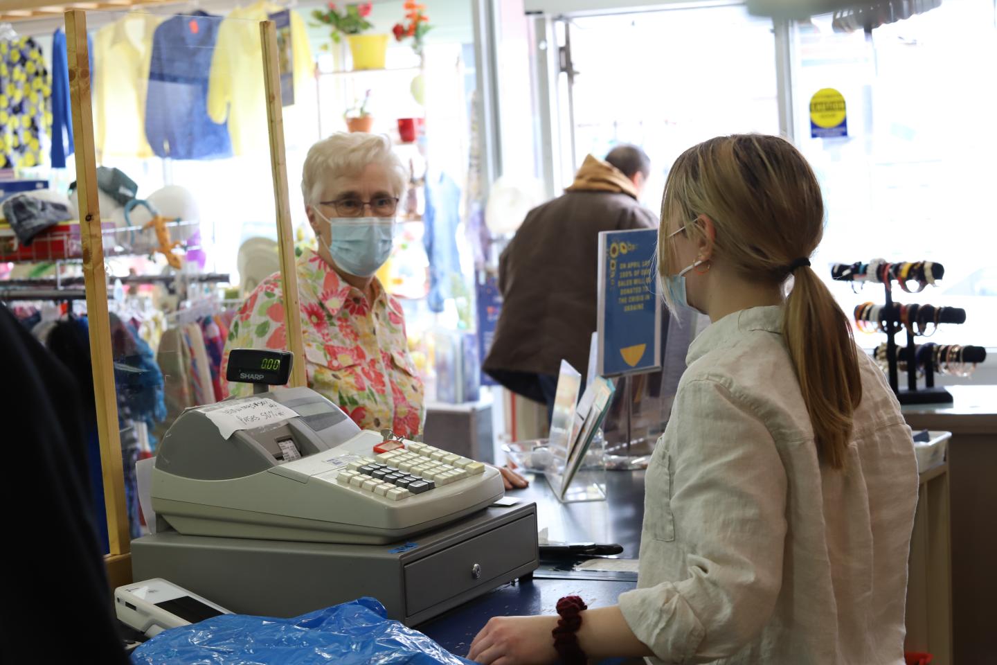 An older woman in a floral shirt and face masks stands in front of a cashier counter at a thrift shop.