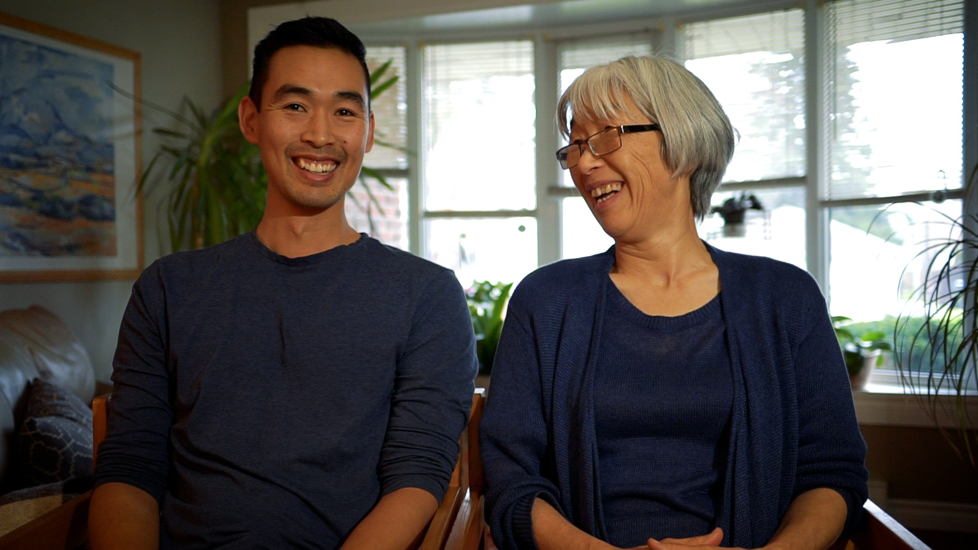 An older mother and son sit next to each other. The son is looking at the camera. The mother is looking at the son.