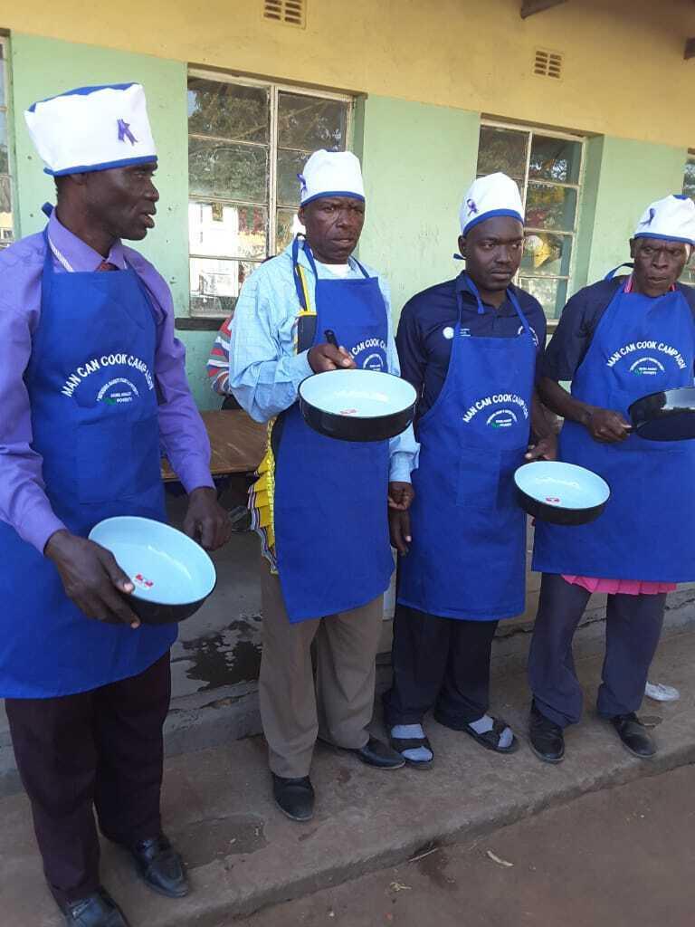 A group of four men in blue aprons and white caps holding skillets