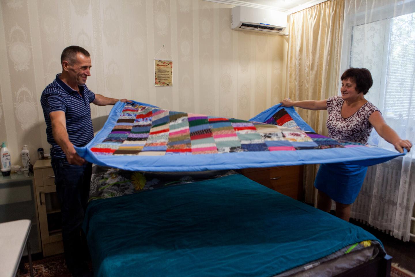 A Ukrainian man and woman work together to put a colorful comforter on a bed