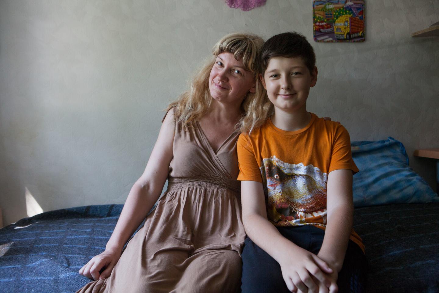 A Ukrainian mother and son sit together on a bed and smile for the camera