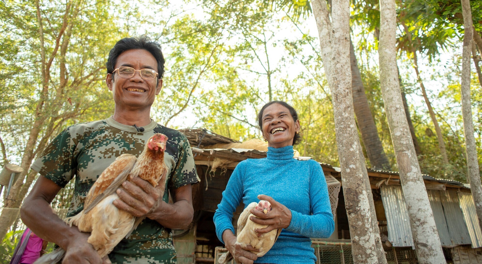 A Cambodian man and woman each hold a chicken. The woman is laughing.