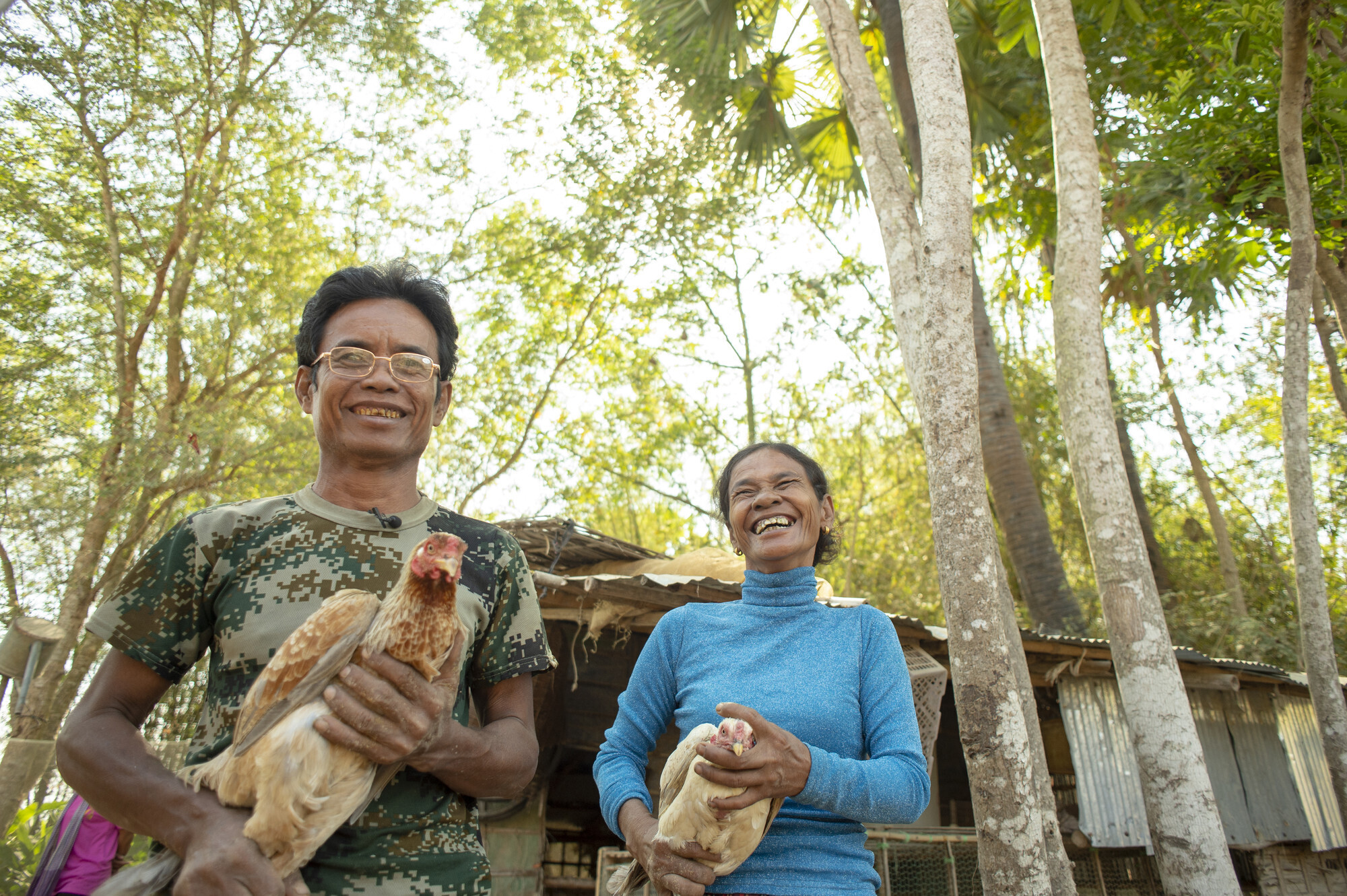 A Cambodian man and woman each hold a chicken. The woman is laughing.