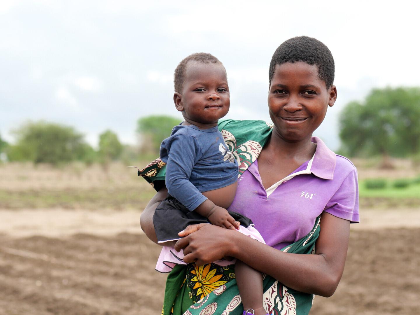 A mother holds her child. She is standing in a field.