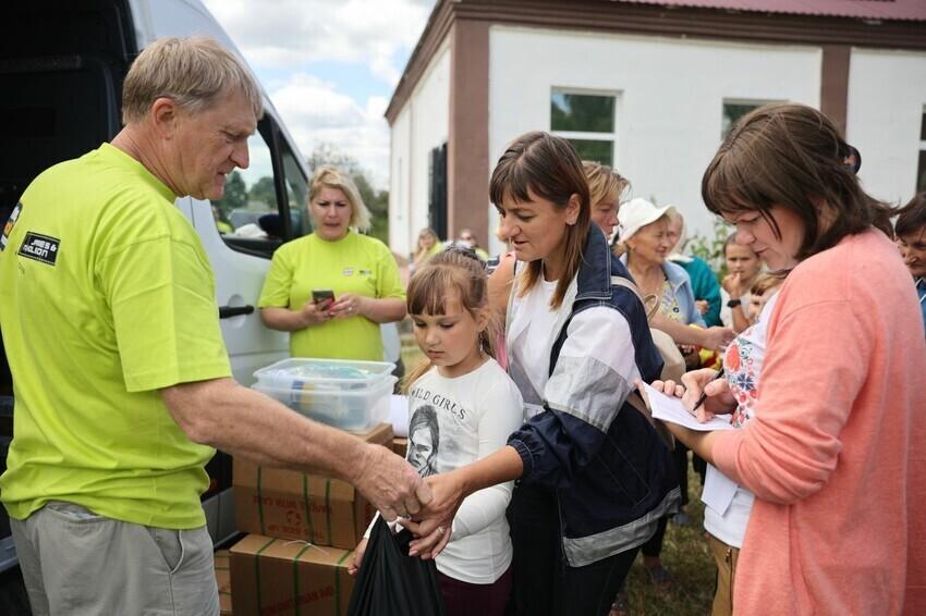 A Ukrainian mother and daughter receive a bag of humanitarian aids from a volunteer in a neon shirt. Another volunteer stands to the right with a clipboard.
