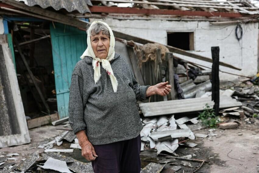 An elderly Ukrainian woman with a scarf tied around her head walks in the wreckage of her destroyed home.