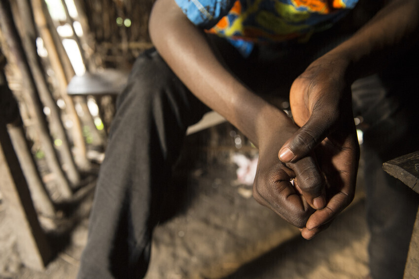 A close-up photo of clasped hands. The photo is taken from an eagle-eye view above the person, with a dirt floor below their hands.