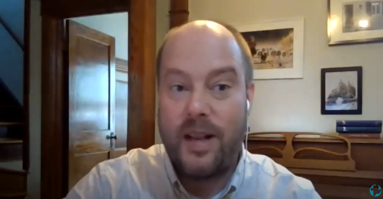A balding man with beard and a white collared shirt speaks into camera during video call